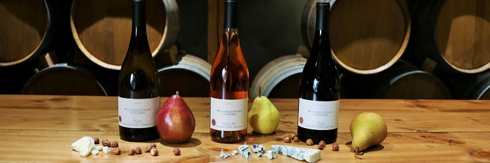 Three bottles of WVV wine with pears, cheese and hazelnuts on the table with barrels behind