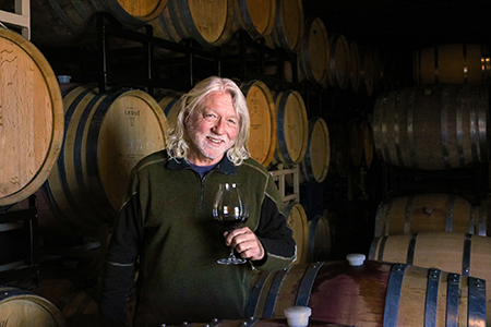 Winemaker, Terry Culton standing in front a wine barrels while holding a glass of Pinot Noir close to his face as we prepares to take a drink.