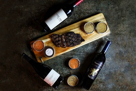 Image of a number of red wines surrounding a wooden cutting board featuring BBQ.