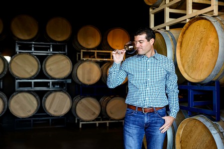 Winemaker, Greg Urmini standing in front a wine barrels while holding a glass of Pinot Noir close to his face as we prepares to take a drink.