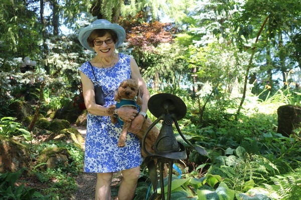 A woman in a blue floral dress and hat holds a small dog in a lush garden setting.