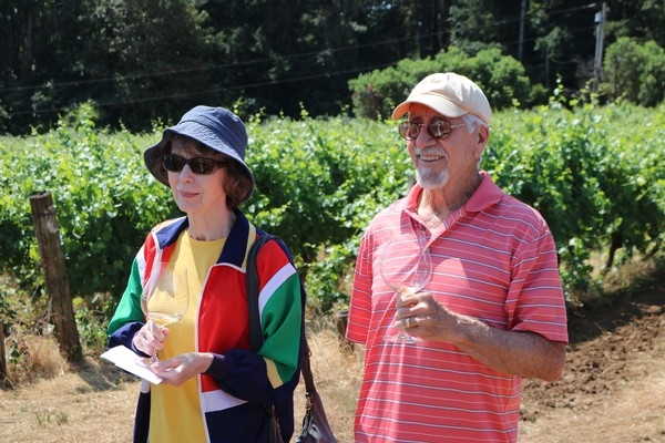 A woman in a colorful jacket and hat stands with a man in a striped shirt, both holding glasses, in a vineyard setting.